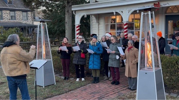 A group of people singing Christmas carols outdoors at night, standing in front of a house with holiday decorations. Two outdoor heaters are visible, one on each side, warming the scene.