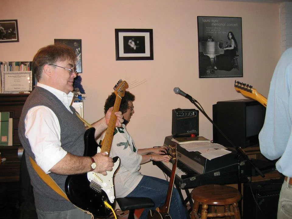 Musicians playing instruments during a performance in a cozy room with framed pictures and a DVD cover on the wall.