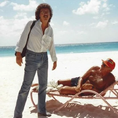 A woman standing on a white sandy beach next to a shirtless man lying on a beach lounge chair, with clear blue waters and a partly cloudy sky in the background.