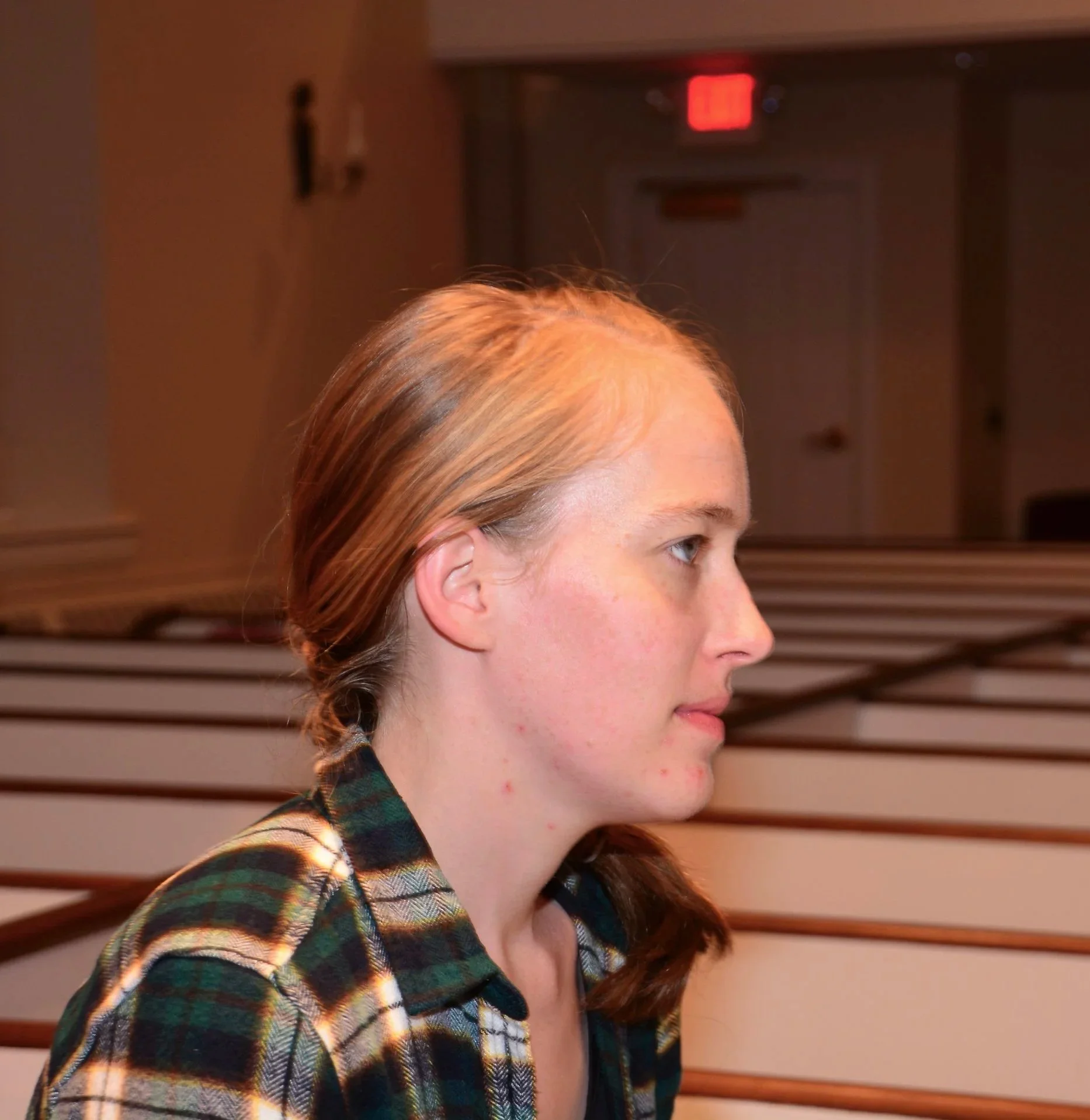 A young woman with red hair and freckles wearing a plaid shirt, shown in profile, facing right, in an indoor setting with wooden slats in the background.