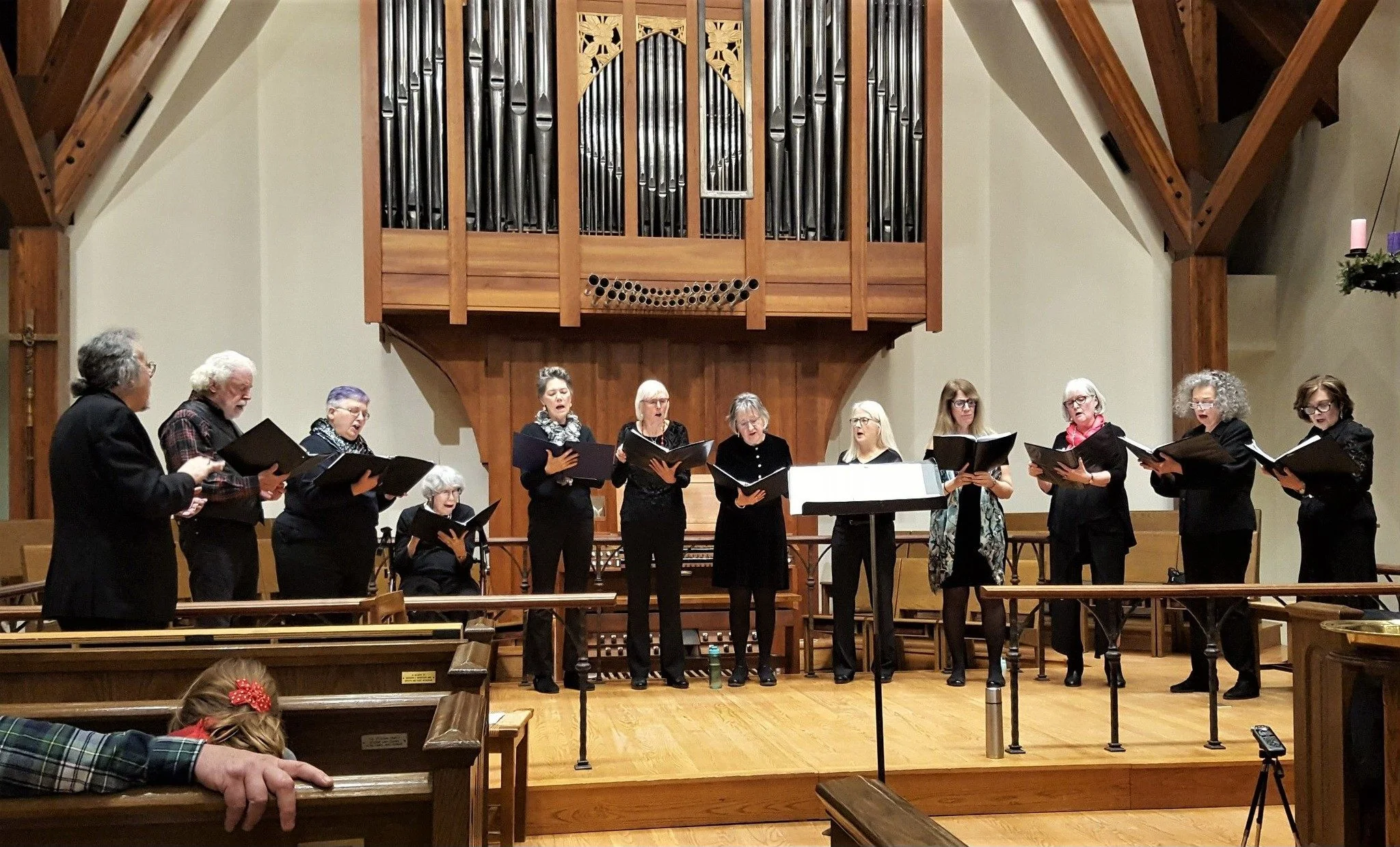 A choir of women and a man singing on a church stage in front of a large pipe organ.