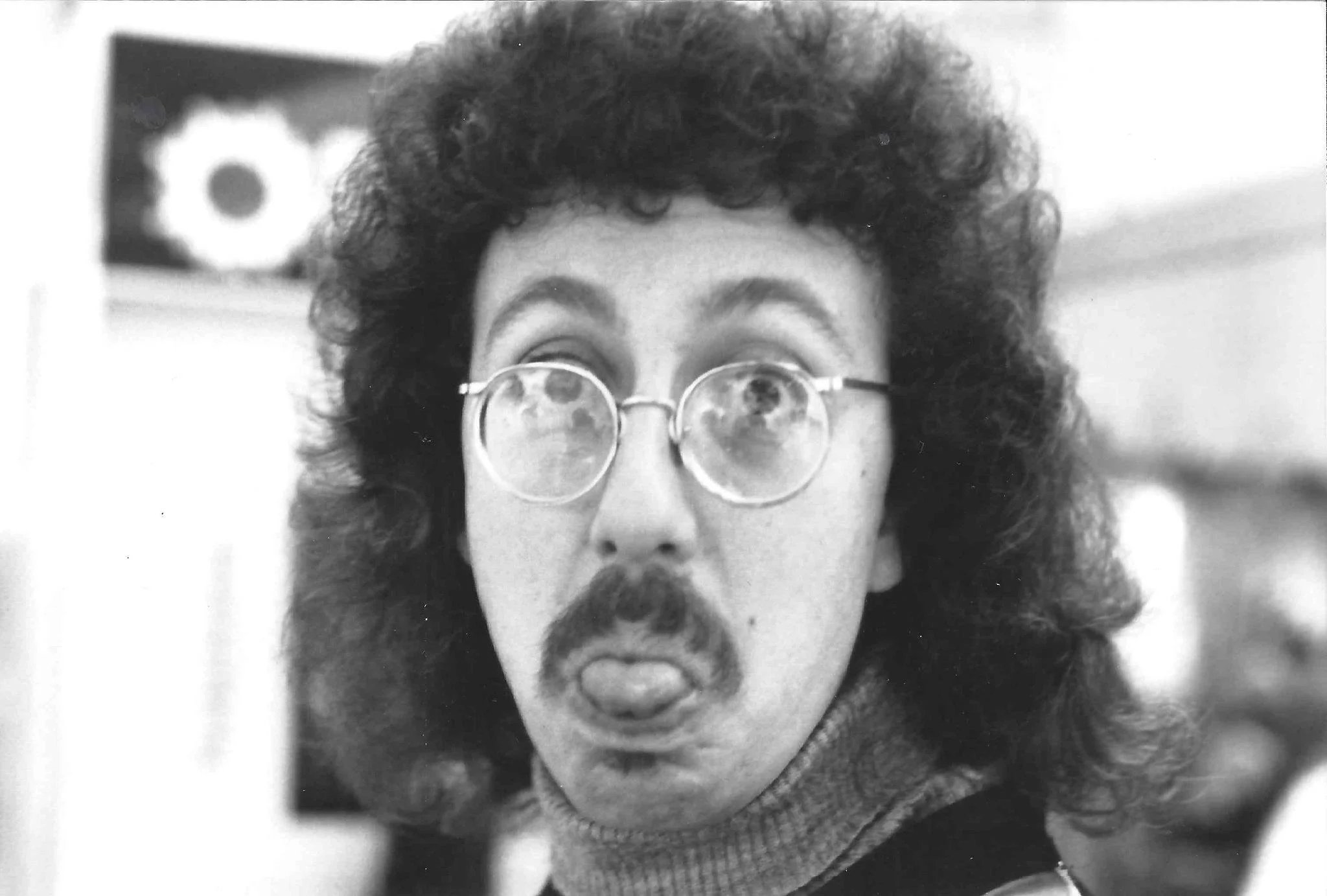 A black-and-white photo of a man with curly hair, wearing round glasses, with a mustache and beard. He appears to be making a playful or exaggerated facial expression, with his tongue slightly out. He is dressed in a sweater and is indoors, with a bl