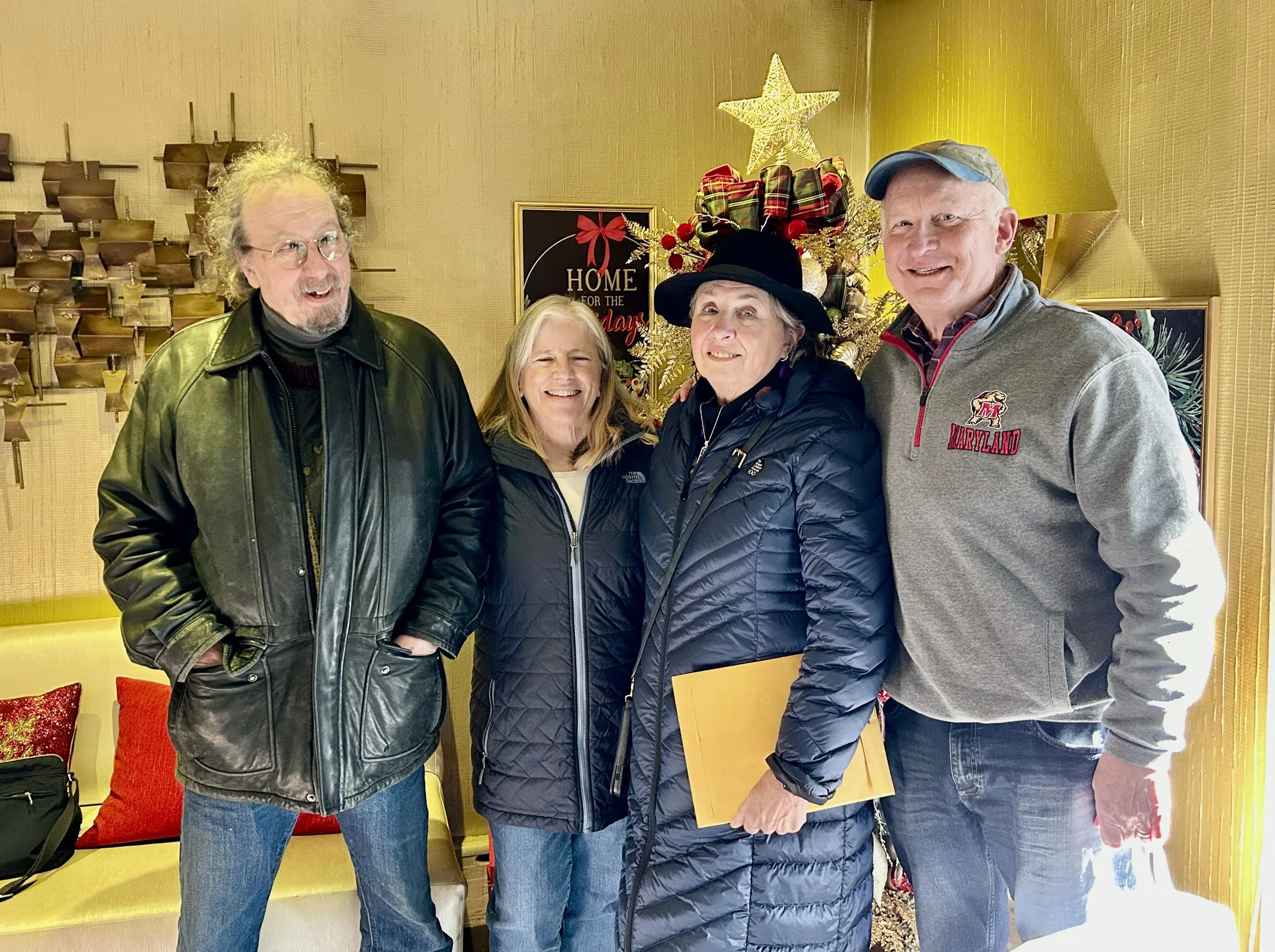 Four smiling adults standing together indoors with holiday decorations, including a Christmas tree with gifts on top and a sign that reads 'Home for the Holidays.'
