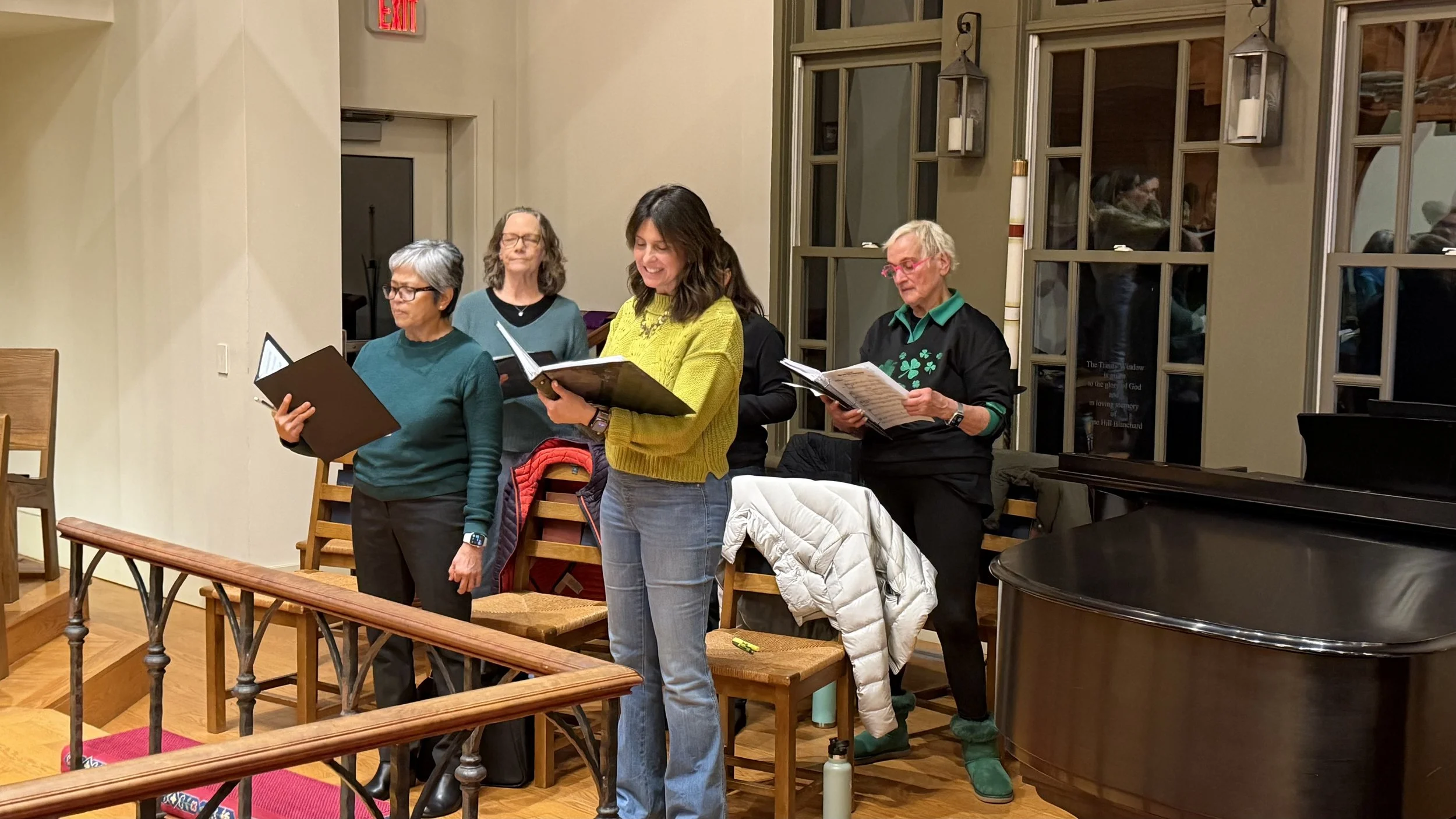 Four women and one man standing in a room, holding sheet music. They appear to be singing or practicing together. The room has large windows, a piano, and chairs, with warm lighting and wooden accents.