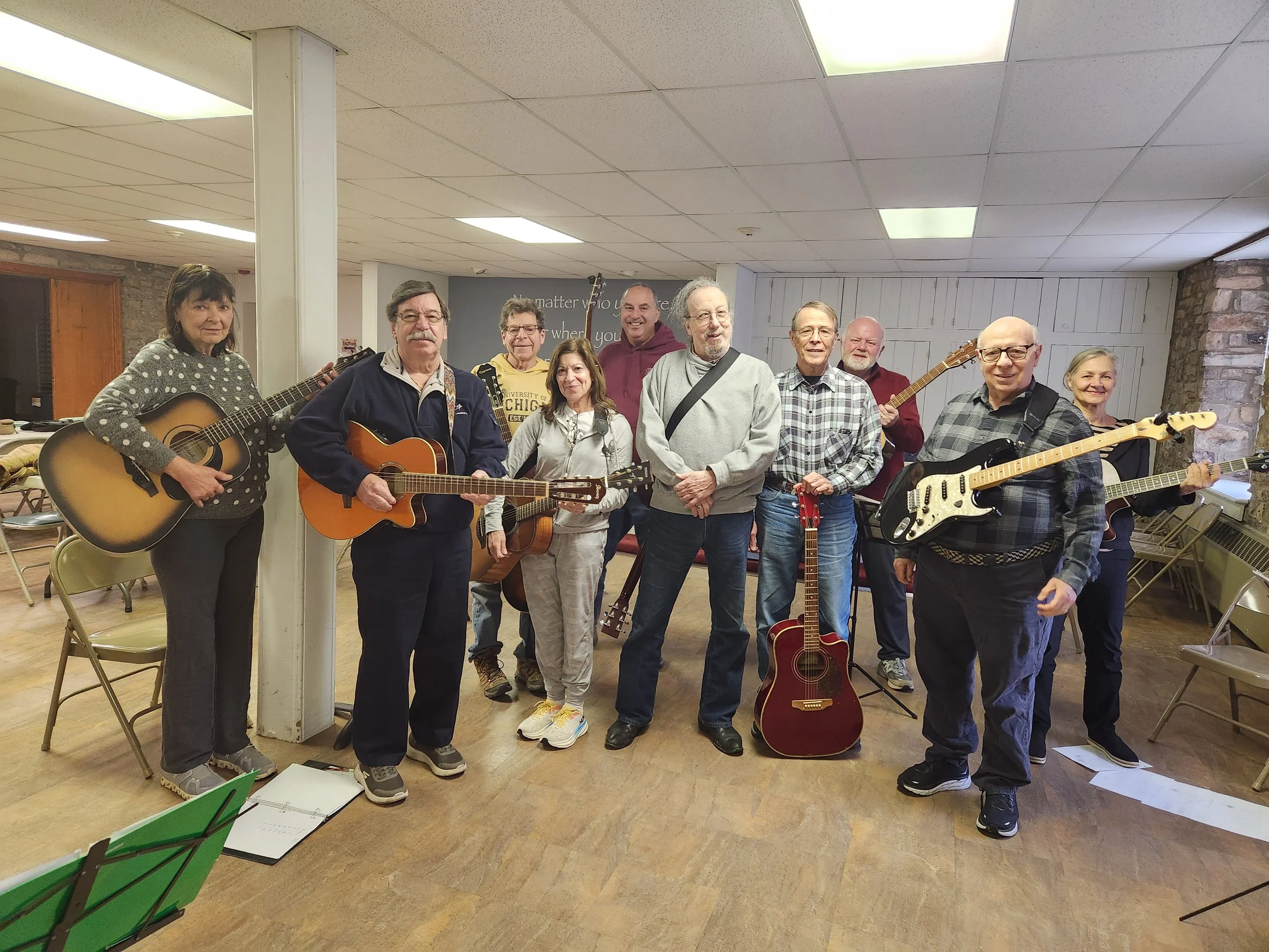 Group of ten people, some holding guitars, standing in a room with a stone wall and white shiplap, posing for a photo.
