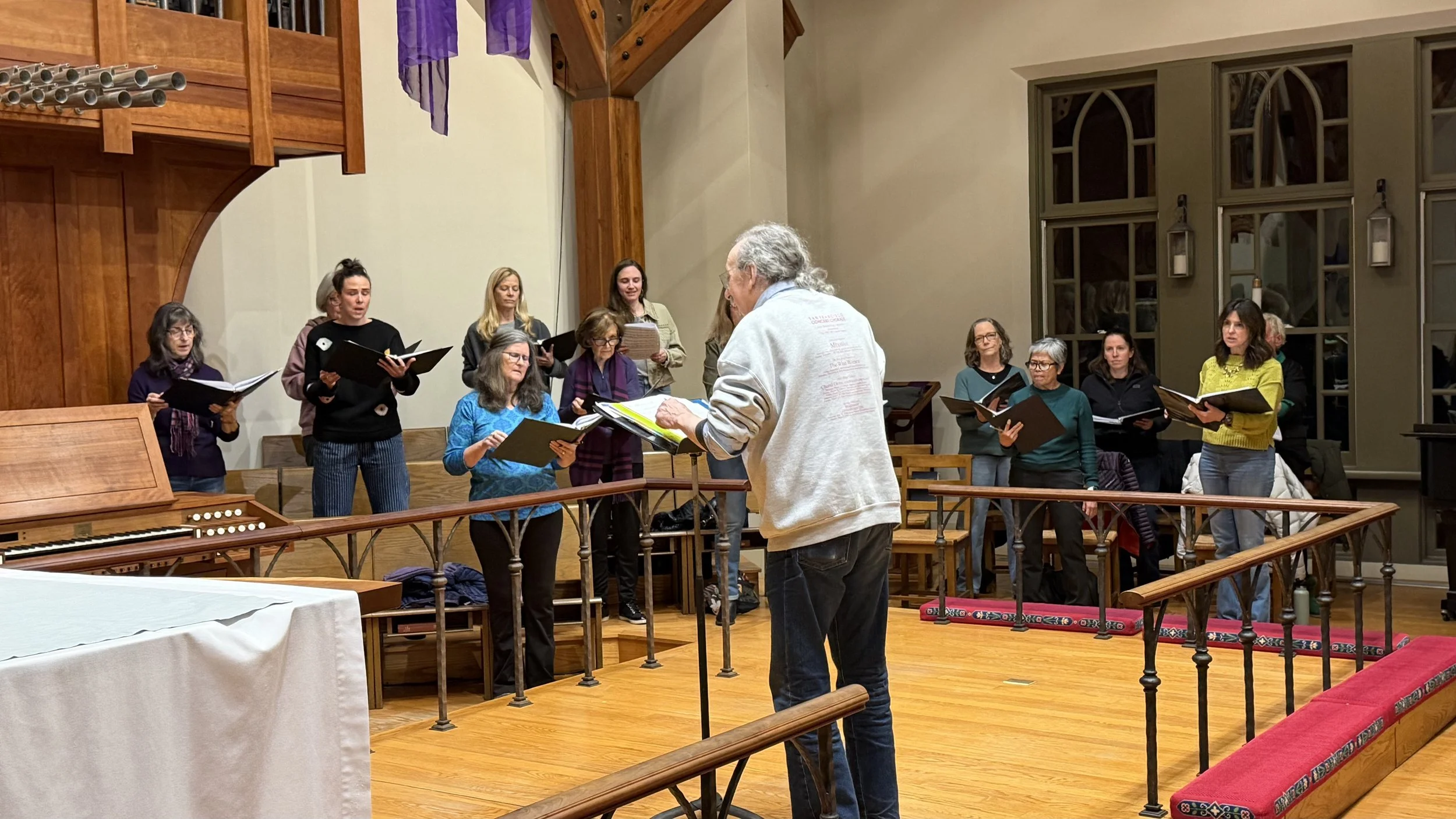 A choir of women holding sheet music practicing with a conductor in a church or concert hall.
