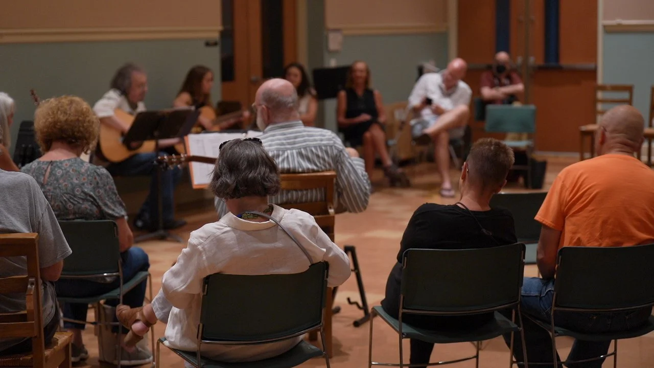 A group of people sitting in a semi-circle, listening to a musical performance or a talk in a cozy, wood-floored room with green and beige walls.