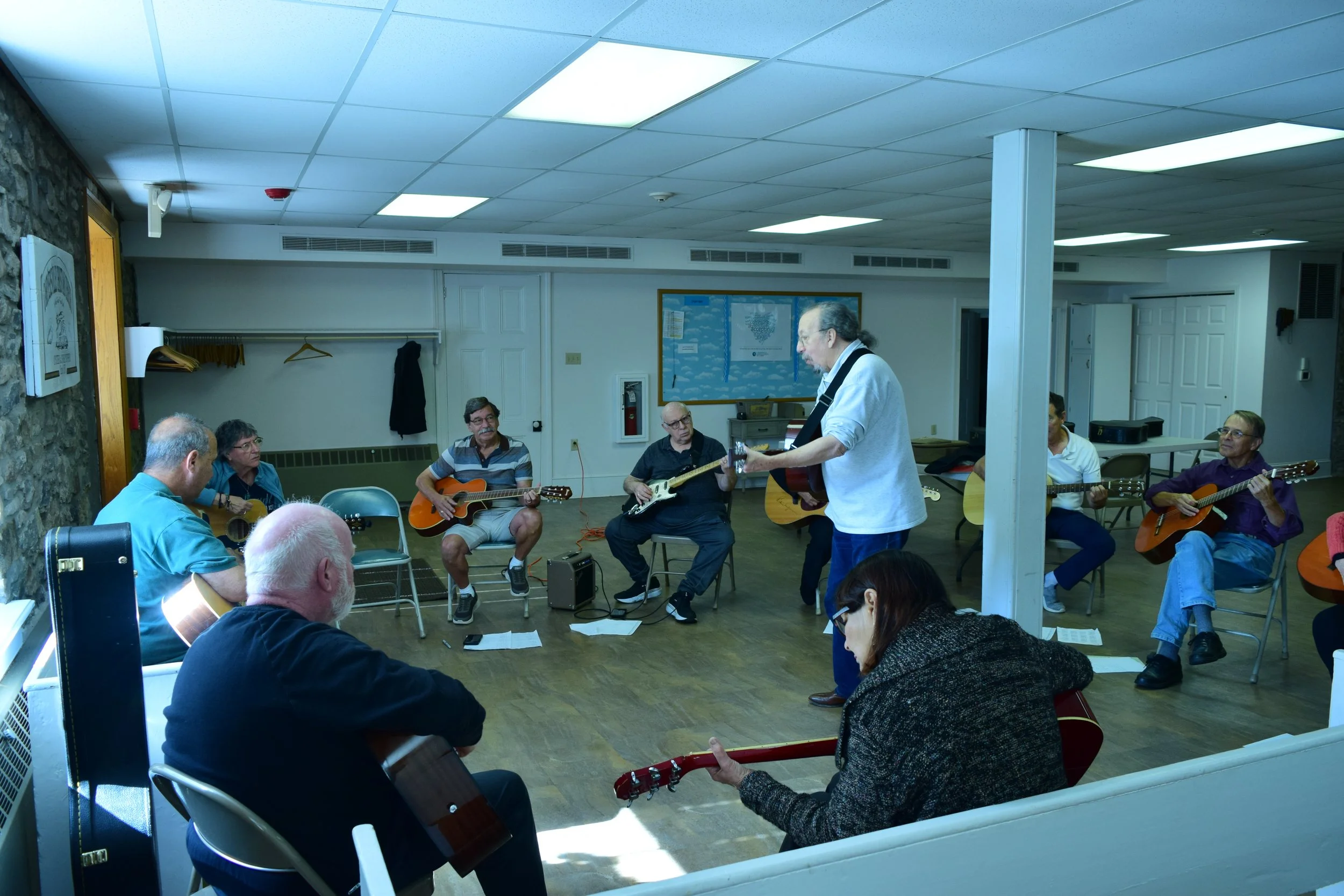 Group of people seated in a circle, playing guitars and singing in a room with white ceiling tiles and a stone wall. A person stands in the center, possibly leading the activity.