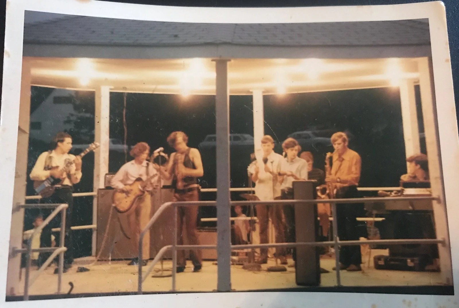 A group of musicians performing on an outdoor stage at night, with some playing guitars and others singing