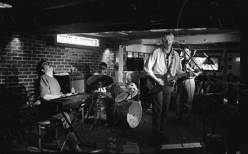 A black and white photo of a live music band performing in a brick-walled venue. The band includes a keyboard player, drummer, and two guitarists, one of whom is singing into a microphone.
