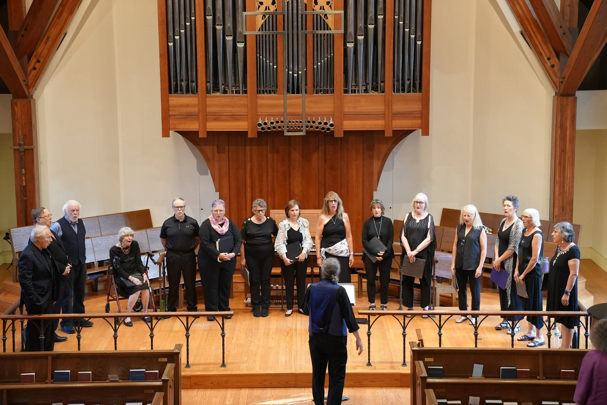 A group of people standing on a church altar in front of a wooden pipe organ, apparently preparing for a choir performance, with some holding sheet music or folders, in an interior with wooden accents.