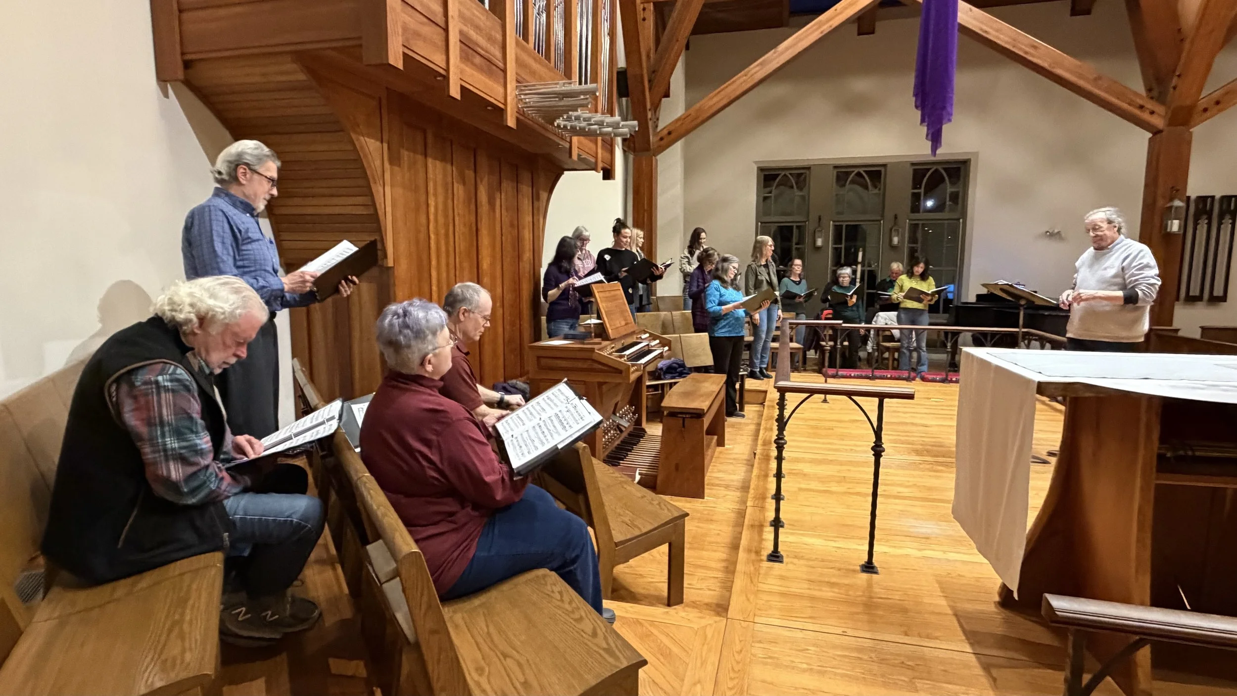 People gathered in a church or chapel listening to a conductor. Some are seated playing instruments, others standing and singing. Wooden interior with a staircase and stained glass windows in the background.