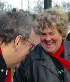 Two people, a man and a woman, smiling and laughing together outdoors. The woman has curly hair, glasses, and is wearing a leather jacket with a red scarf and necklace. The man has short hair and glasses.