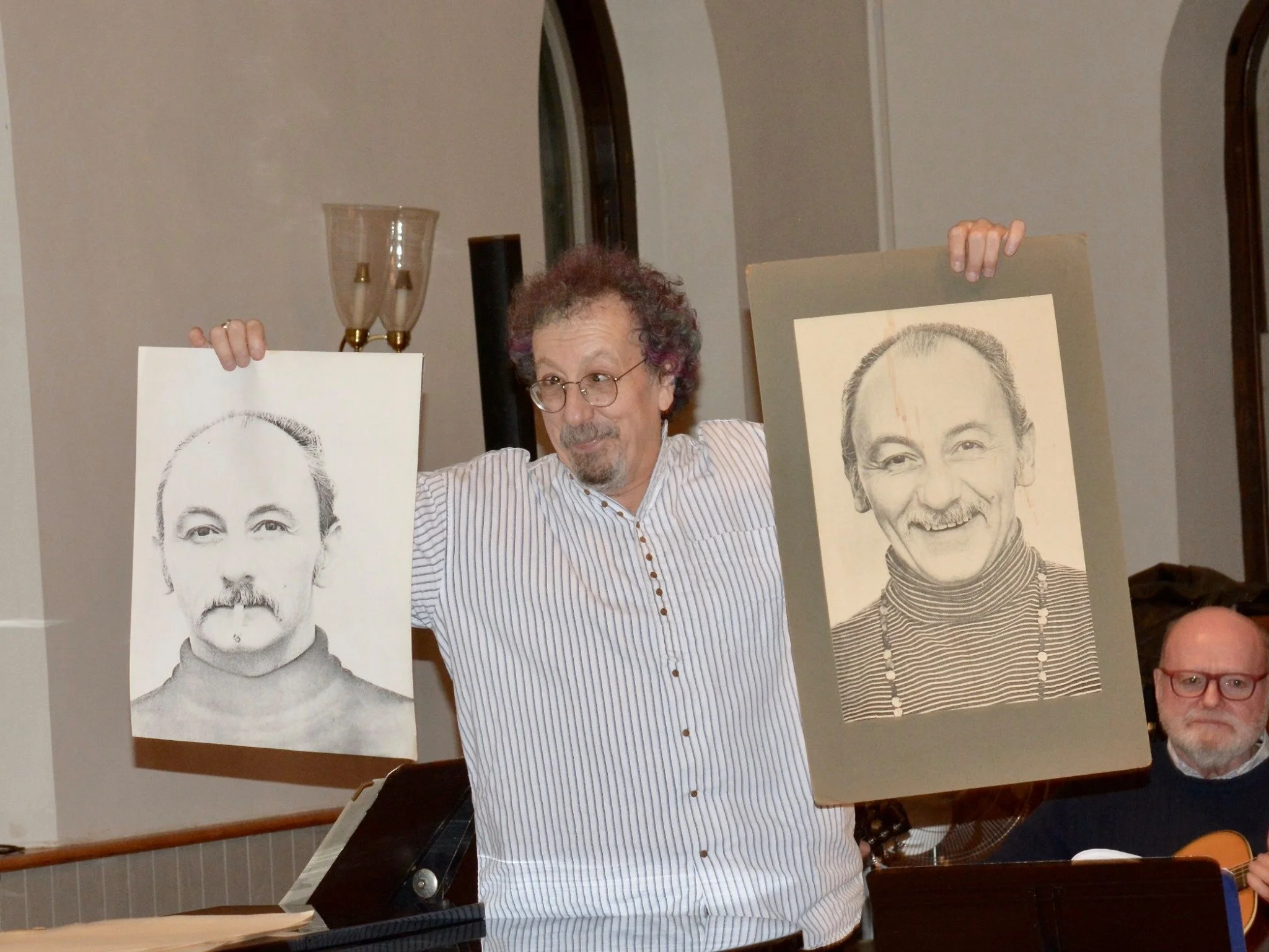 A man with gray curly hair, glasses, and a striped shirt holds up two large portraits of a smiling man with short gray hair, wearing a high-collared shirt. The portraits are black and white sketches or prints.