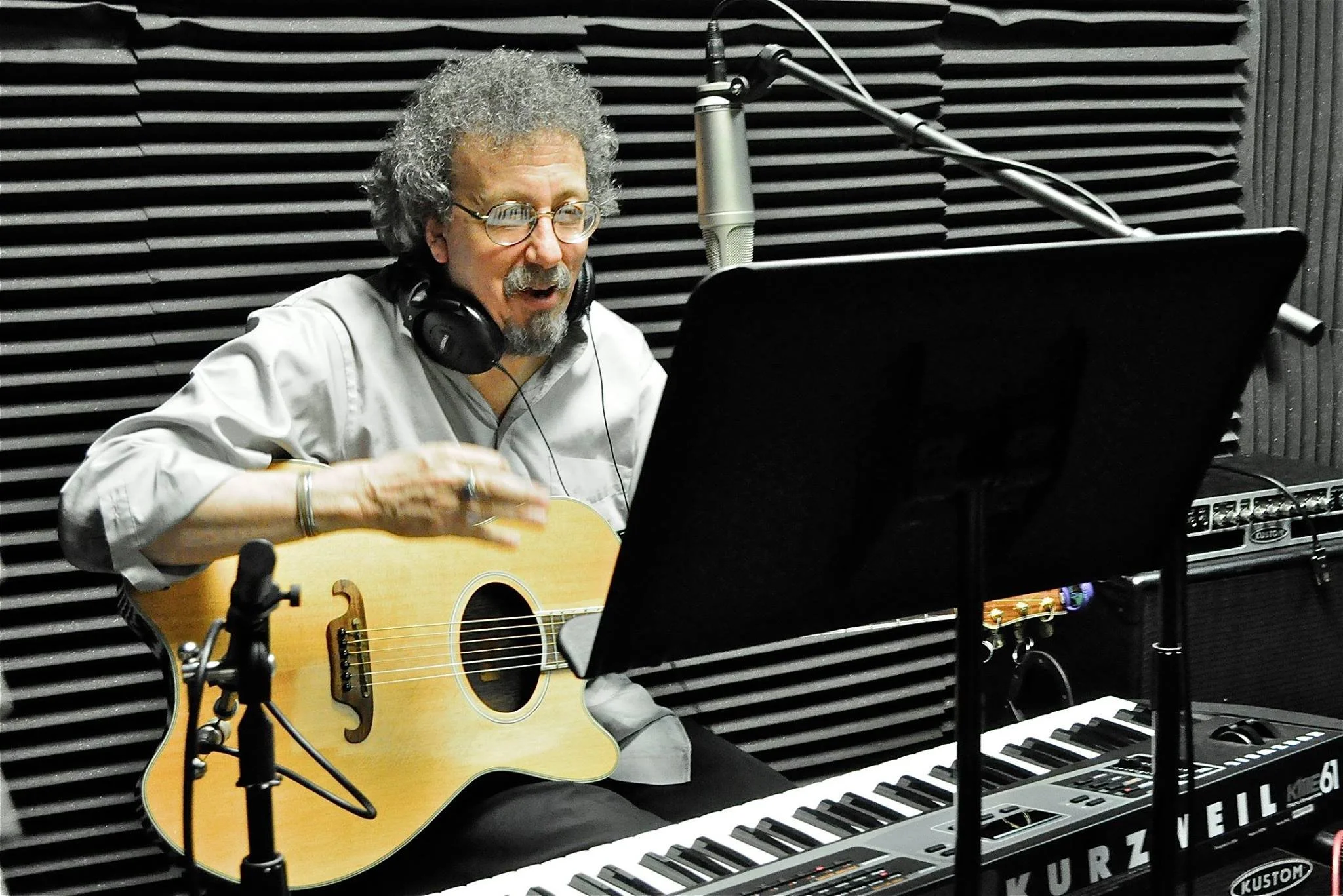 A man with curly gray hair and glasses singing into a microphone while playing an acoustic guitar in a music recording studio, surrounded by musical equipment including a keyboard and a music stand.