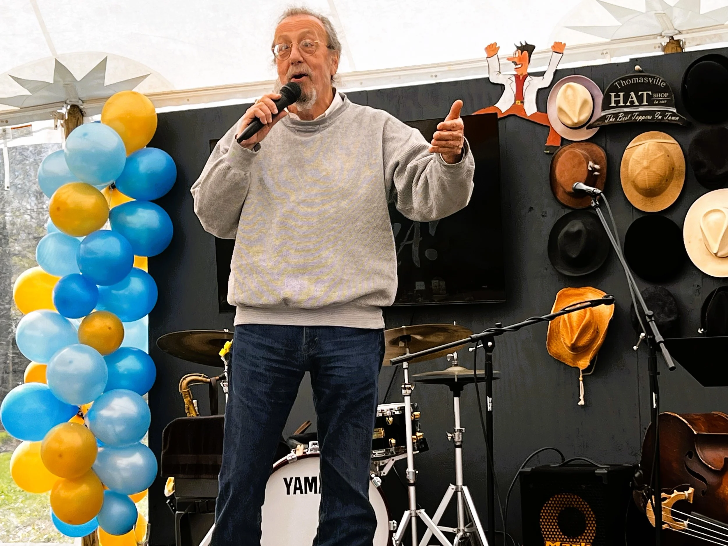 An elderly man with glasses and a gray sweatshirt speaking into a microphone at a festive outdoor event. Behind him, there are yellow and blue balloons, a drum set, and a wall decorated with hats and a cartoon figure of a man in a white jacket and re