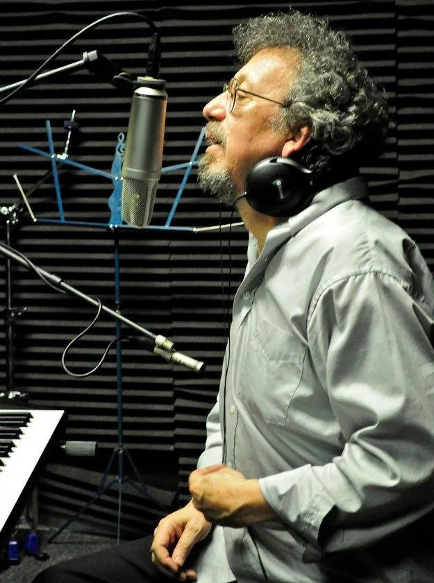 A man with curly gray hair and glasses, wearing a gray shirt, is singing into a large studio microphone while wearing headphones in a recording studio with black soundproofing panels.