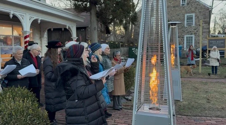 A group of people gathered outdoors during winter, standing in front of a tall, outdoor heater with flames, singing or reading from sheets of paper.