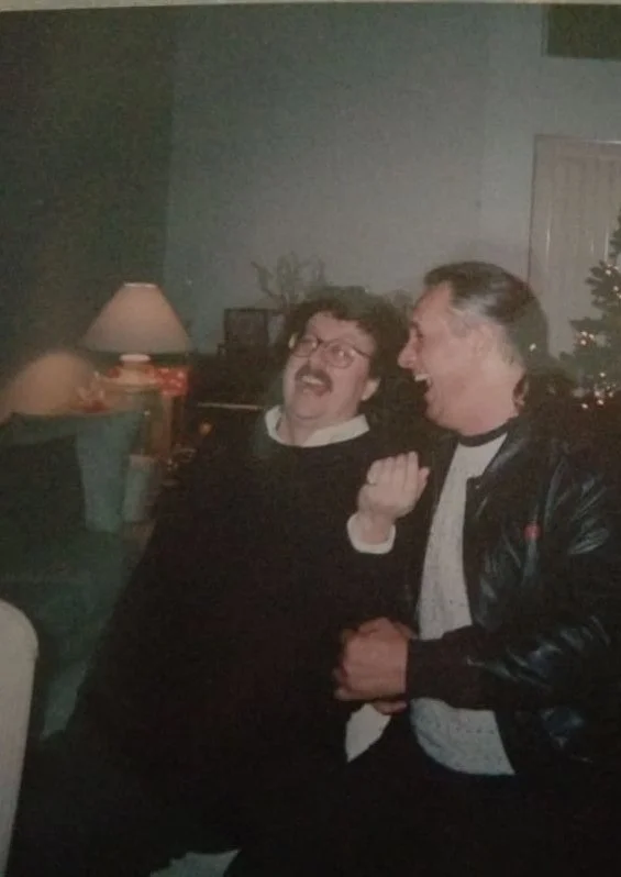 Two men laughing and sharing a joyful moment indoors, with a lit Christmas tree visible in the background.