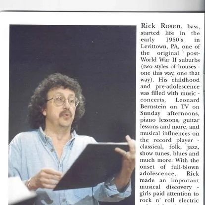 Photograph of Rick Rosen, a man with curly hair and glasses, speaking or performing, with a black folder or book in his hands, against a dark background.