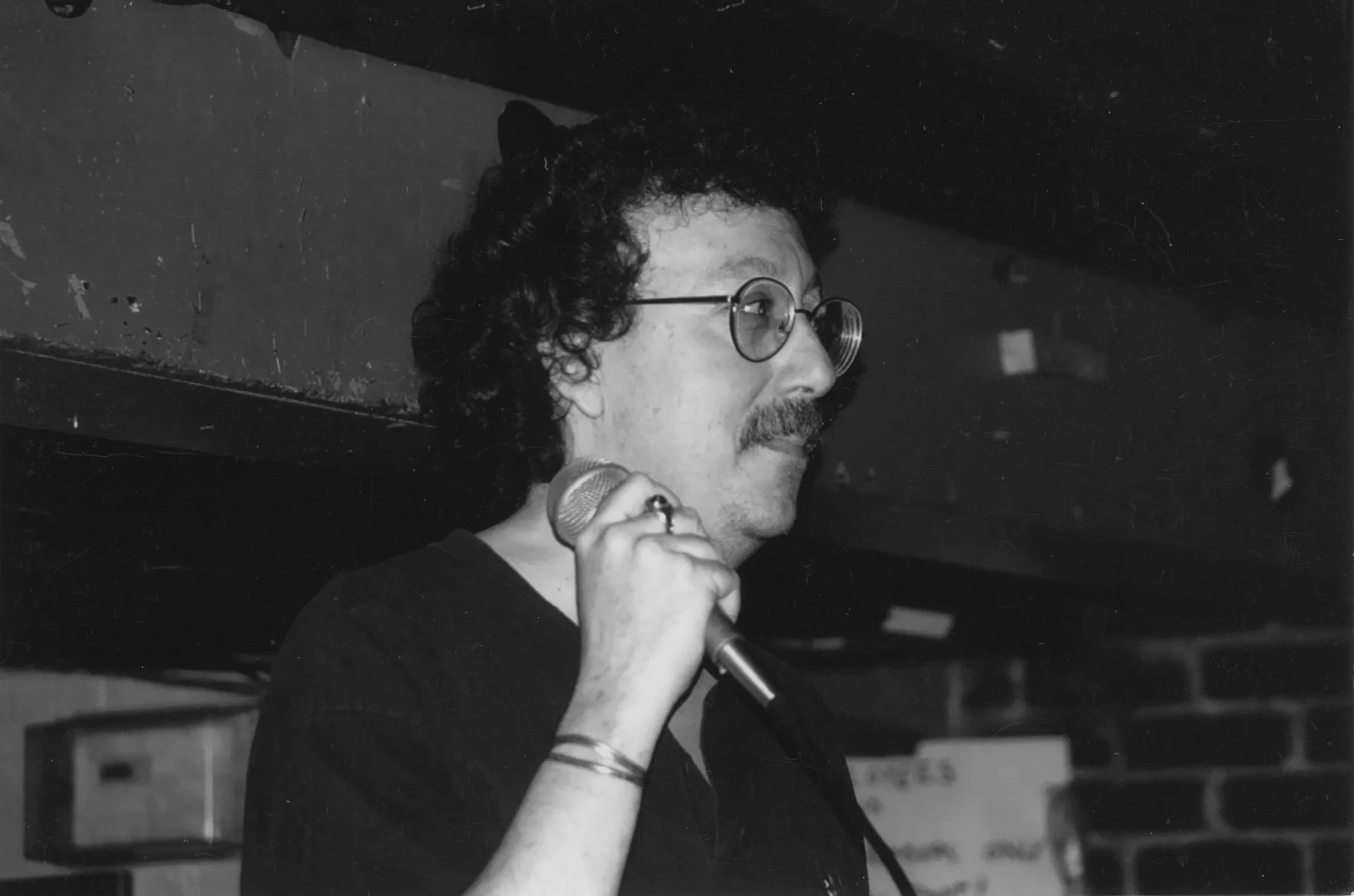 A man with curly hair, glasses, and a mustache is holding a microphone near his mouth, appearing to speak or sing, in a black and white photo.