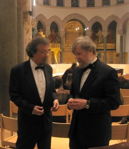 Two men in tuxedos having a conversation in a church or cathedral.