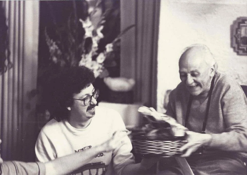 Two men are sitting at a table, both smiling and appearing happy. One is wearing glasses and a light-colored sweatshirt, and the other is bald, wearing a long-sleeve shirt. The man on the right is handing a basket to the man on the left. The backgrou