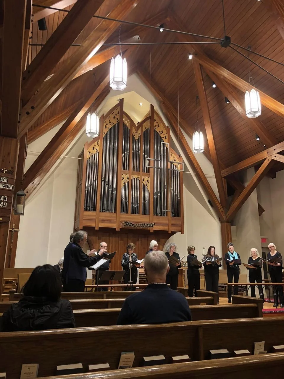 A choir of eleven women and one man performing on stage in a church or concert hall with wooden architecture. The conductor is standing to the side, leading them. The large pipe organ is visible in the background.