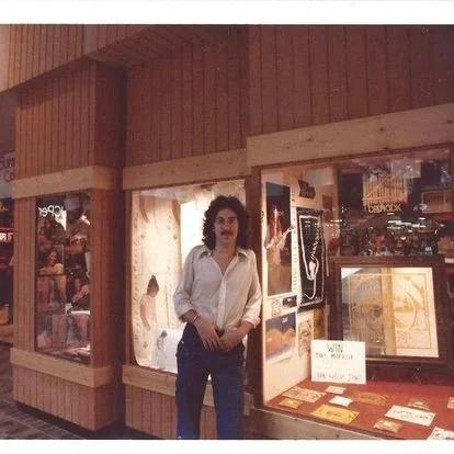 A man with curly hair, wearing a white shirt, standing in front of glass display cases inside a store or museum, with various framed items and memorabilia.