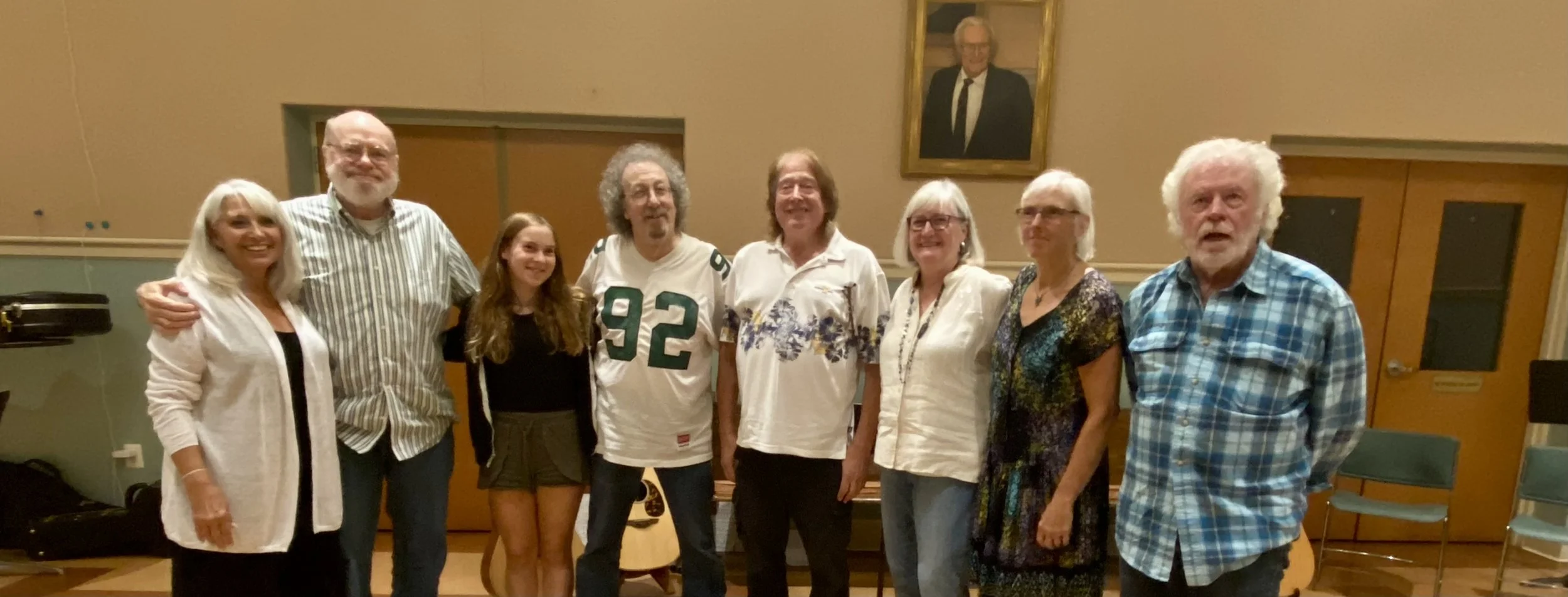 A group of nine people standing together in a room, smiling for a photo. Behind them, a portrait painting hangs on the wall.