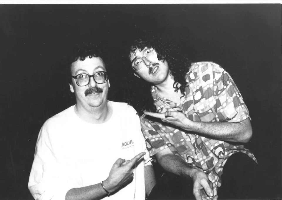 Two men with curly hair and glasses, one with a mustache, posing for a black and white photo, both pointing at the other man.