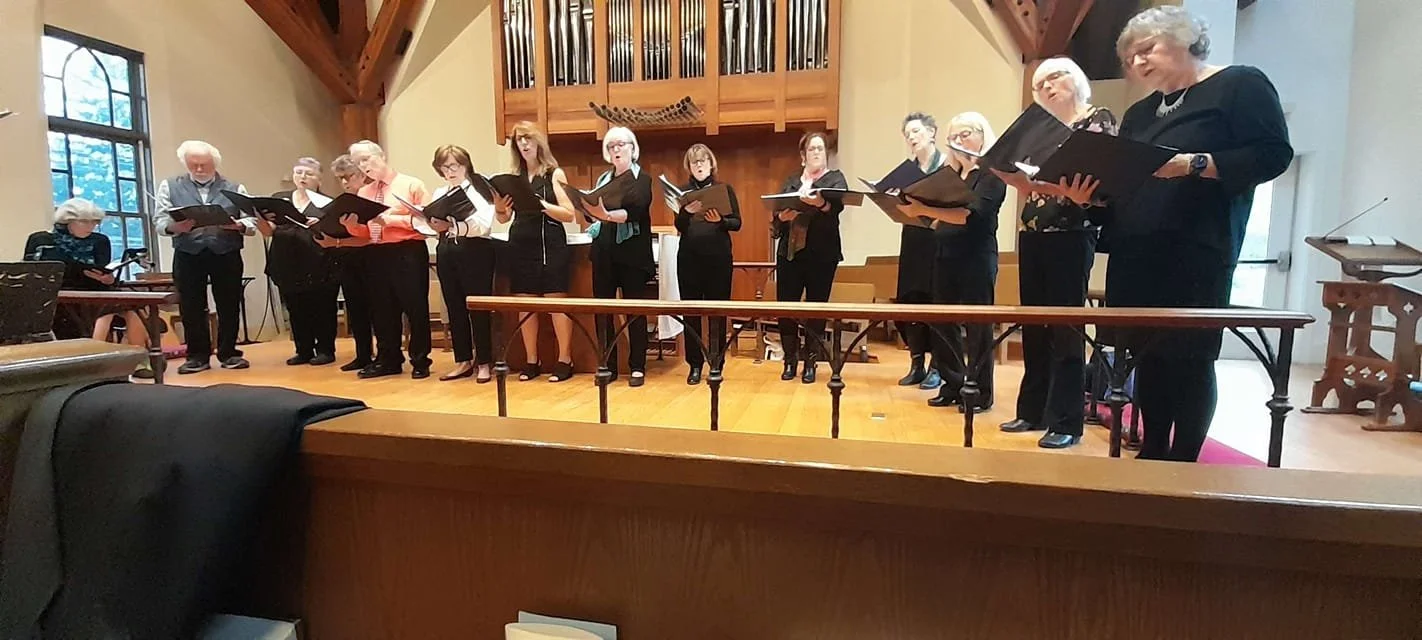 A choir of adult women and one young girl singing on a church stage, holding sheet music, with a wooden organ in the background.