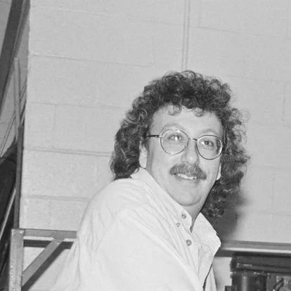 A man with curly hair, glasses, and a mustache smiling, wearing a light-colored shirt, standing indoors.