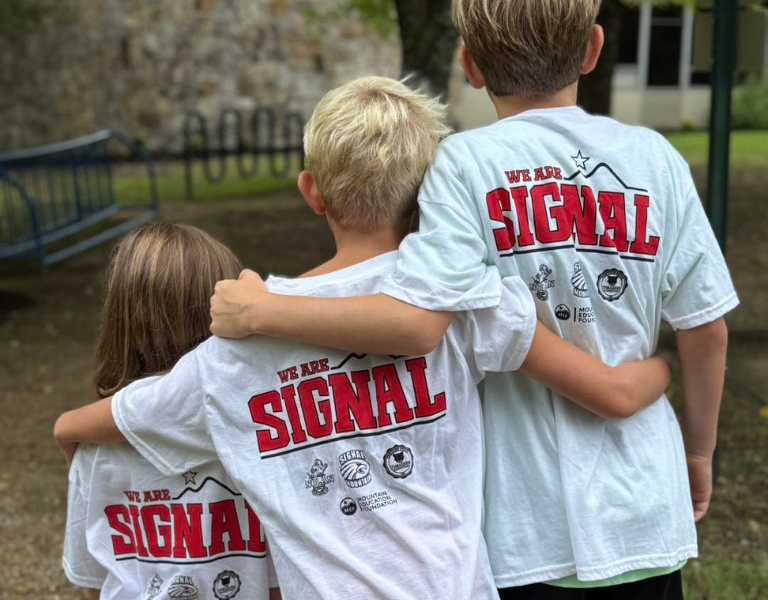 Three elementary aged kids with arms around each other wearing We Are Signal T-Shirts