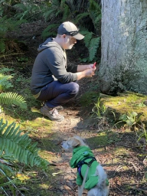 Ian Ameling is crouching in a forest, taking a reference photo with his smartphone, while a small dog in a green jacket looks on.
