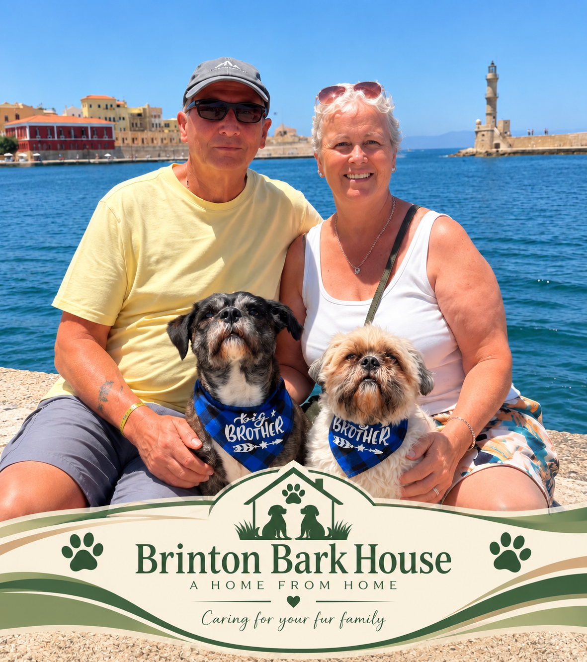 A smiling elderly woman and man sit on a waterfront with two dogs wearing bandanas that say 'big brother' and 'big sister.' They are in front of a sign for Brinton Bark House, with a harbor and lighthouse in the background.