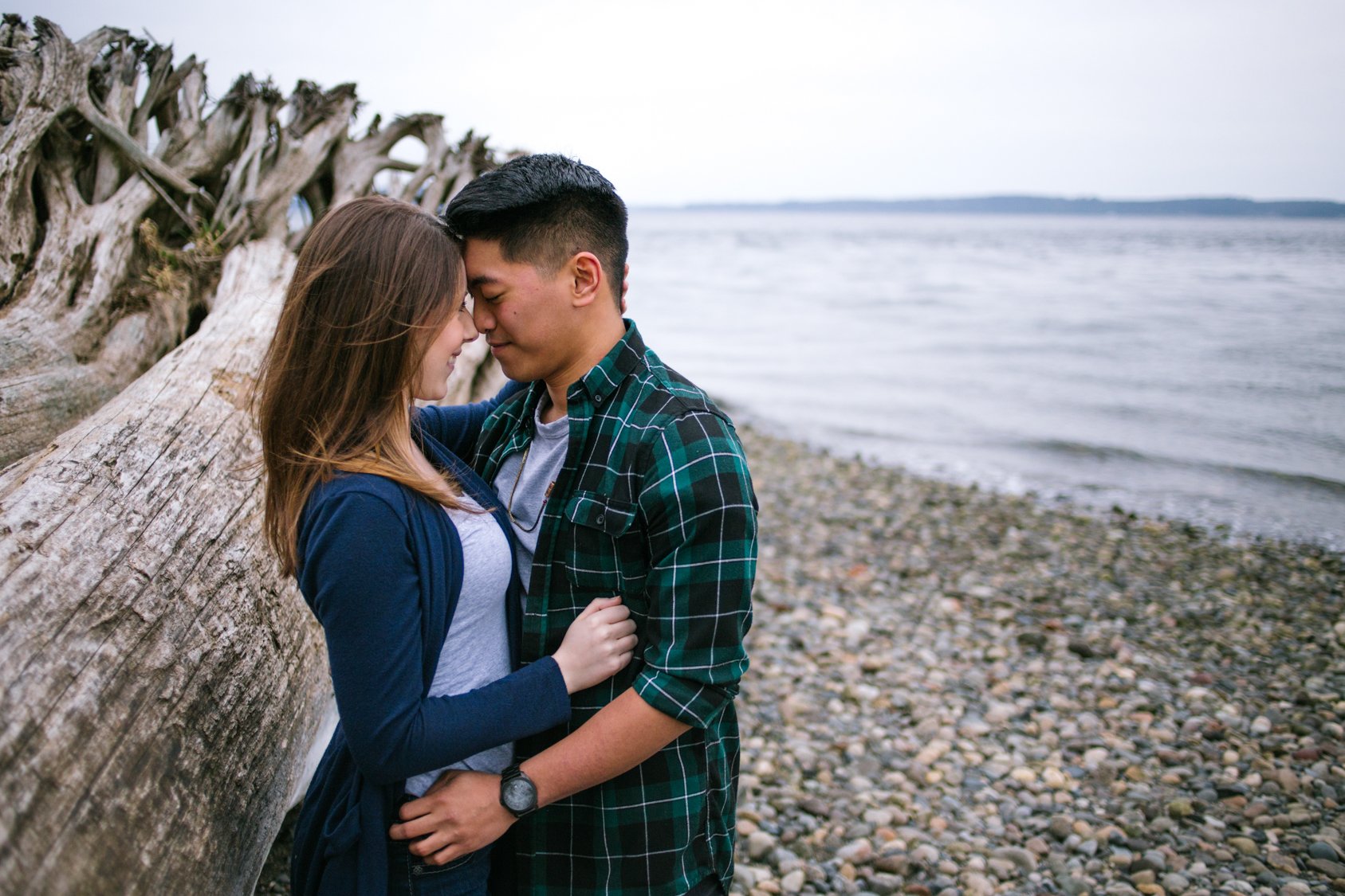 A young couple standing close together by a large driftwood log on a rocky beach, with an ocean and cloudy sky in the background, touching foreheads and smiling.