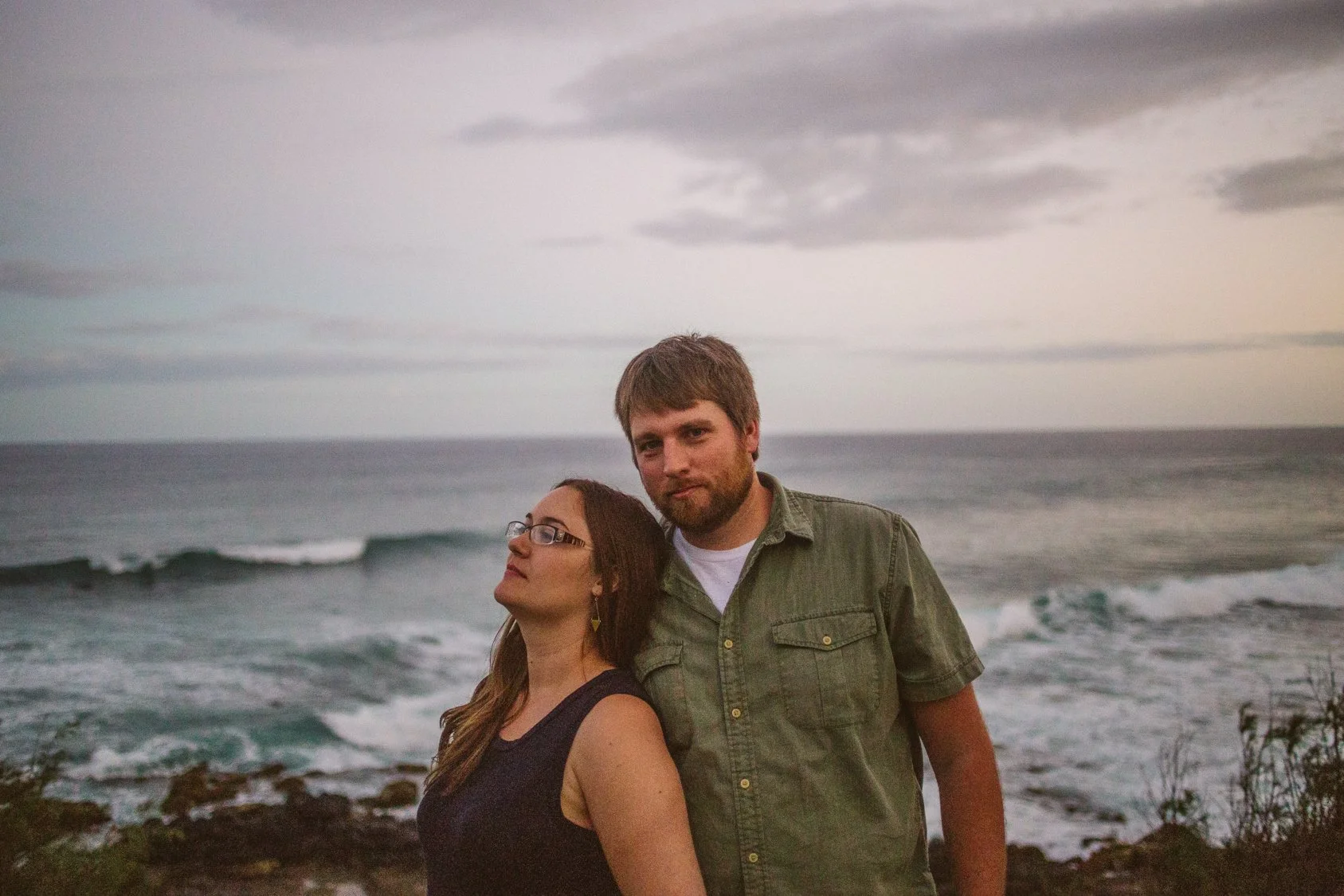 A man and a woman standing close together on a rocky shoreline with the ocean in the background. The woman, wearing glasses and a black tank top, looks slightly upward, while the man, with a beard and wearing a green shirt, looks directly at the came