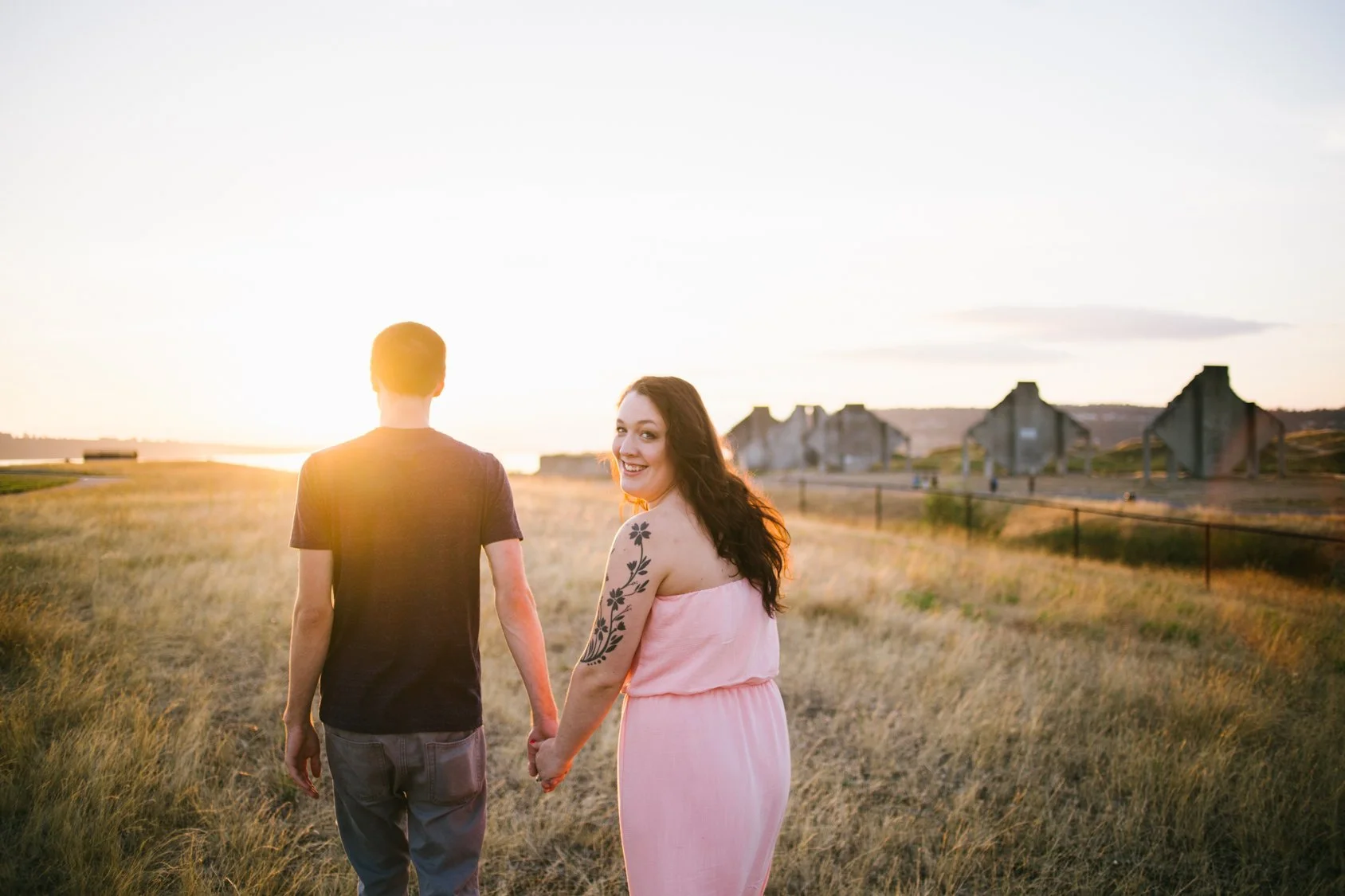 A couple holding hands and walking through a grassy field at sunset, with industrial structures in the background and a smiling woman looking back.
