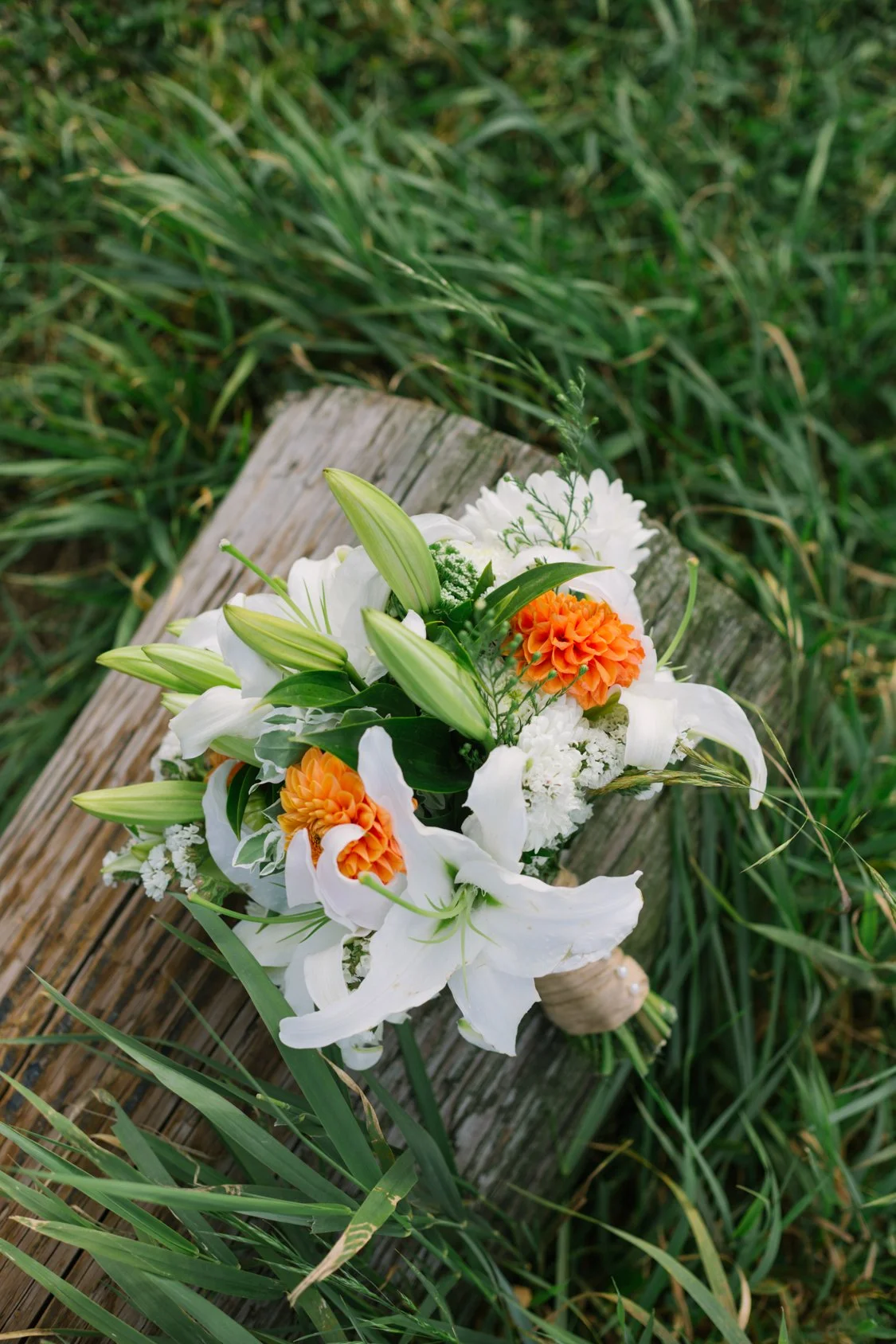 A bouquet of white lilies, orange dahlias, white carnations, and greenery placed on a wooden surface outdoors with grass.