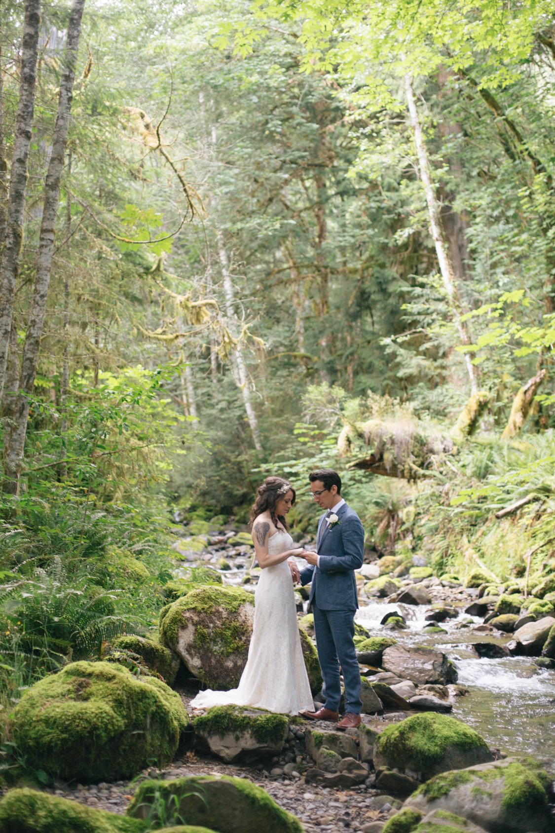 A bride and groom exchanging rings in a lush, green forest with moss-covered rocks and a small stream.