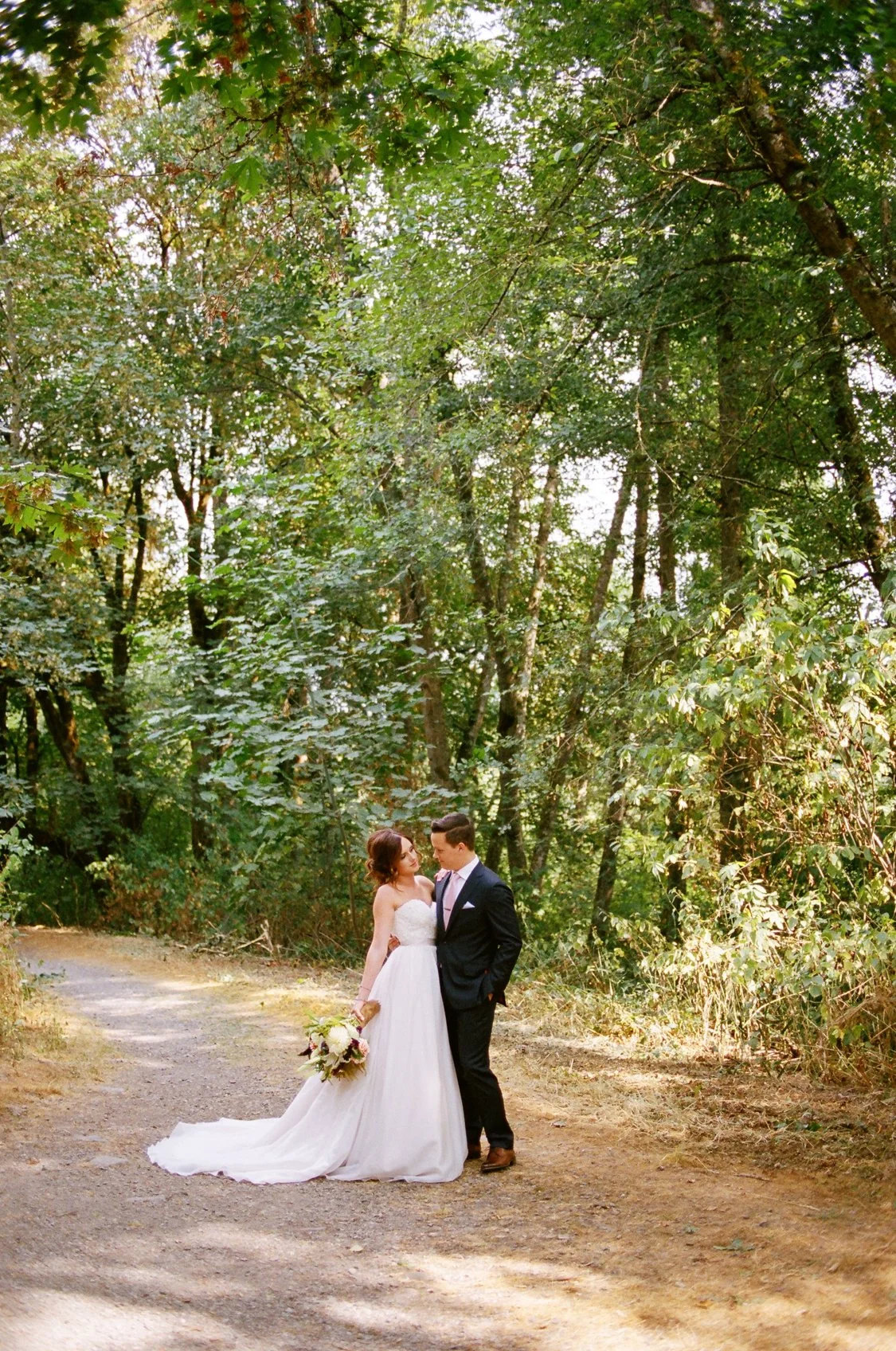 A bride and groom standing on a dirt path surrounded by green trees in a forest, with the bride holding a bouquet and the couple gazing into each other's eyes.