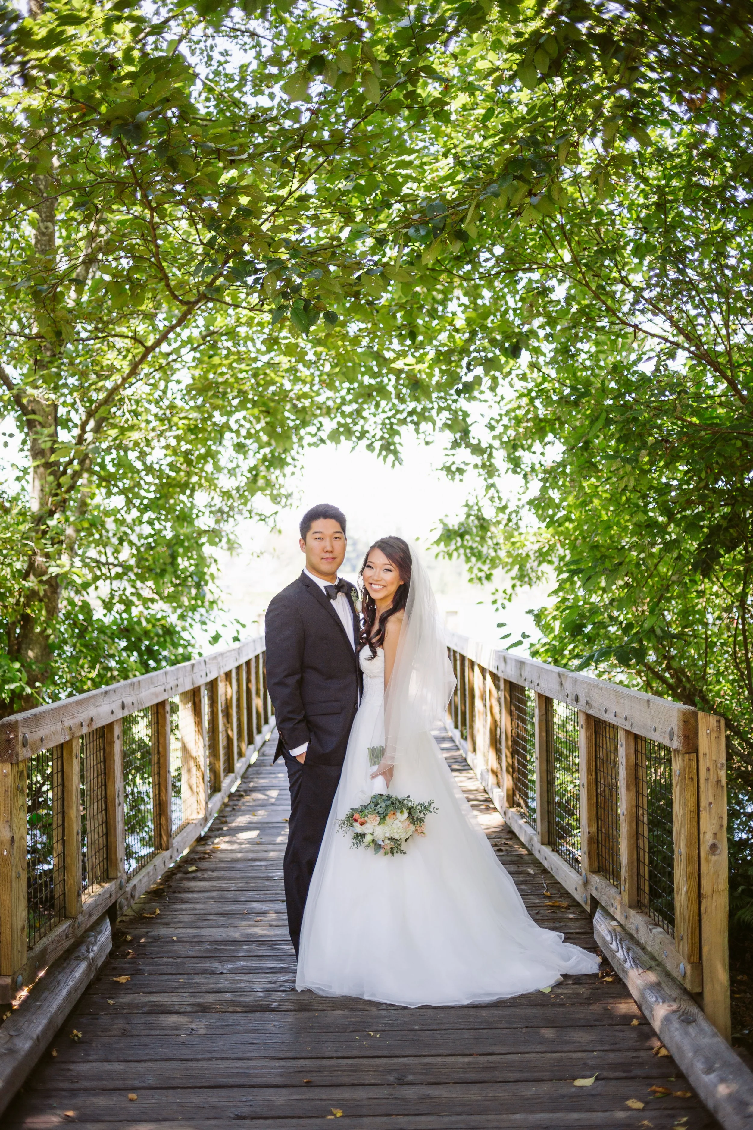 A bride and groom standing together on a wooden bridge surrounded by lush green trees, with the bride in a white wedding gown holding a bouquet and the groom in a black tuxedo. The bride is smiling, and the couple appears happy.