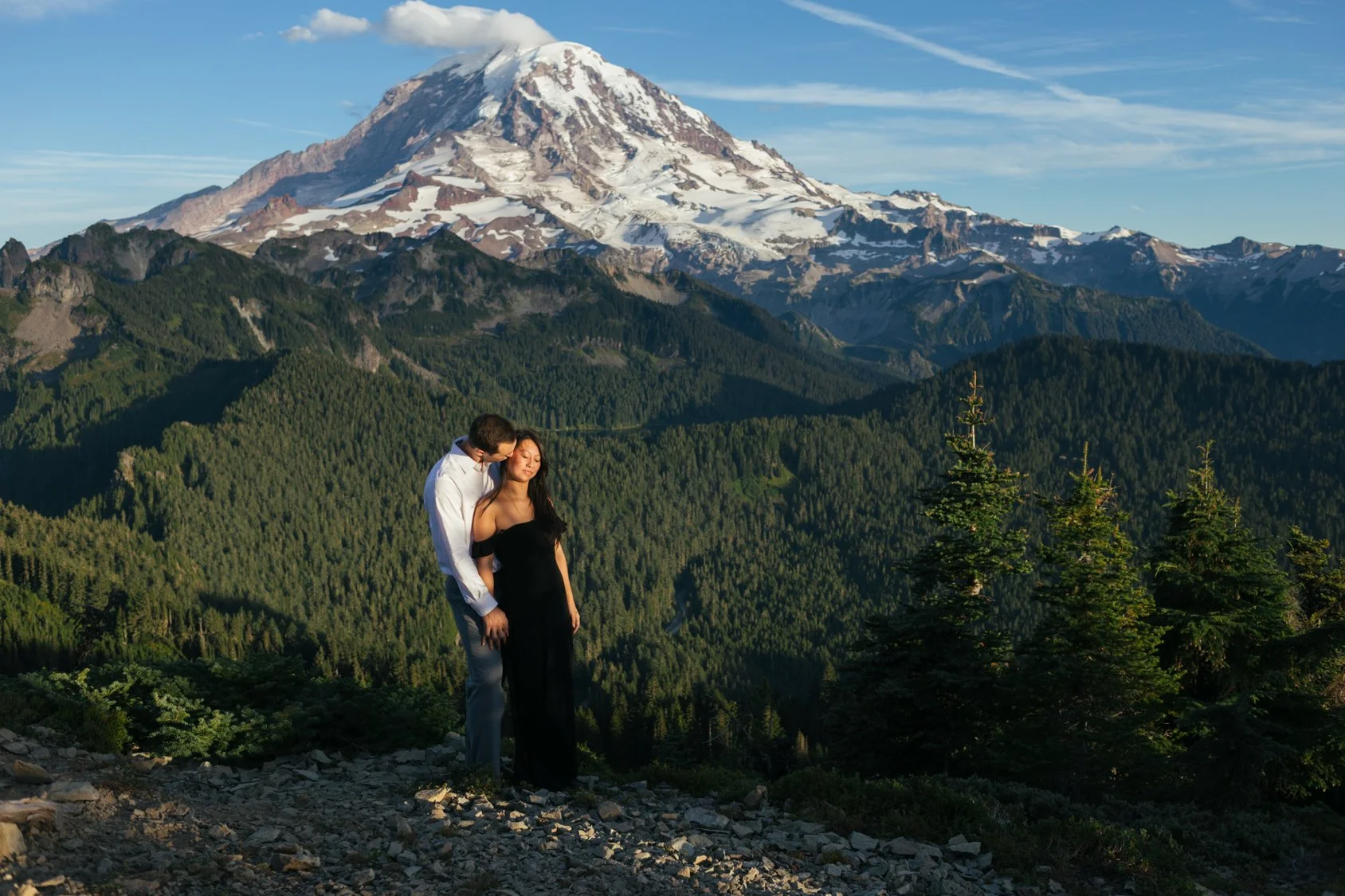A couple stands closely together on a rocky terrain with a backdrop of green forested mountains and a snow-capped mountain peak under a clear blue sky.