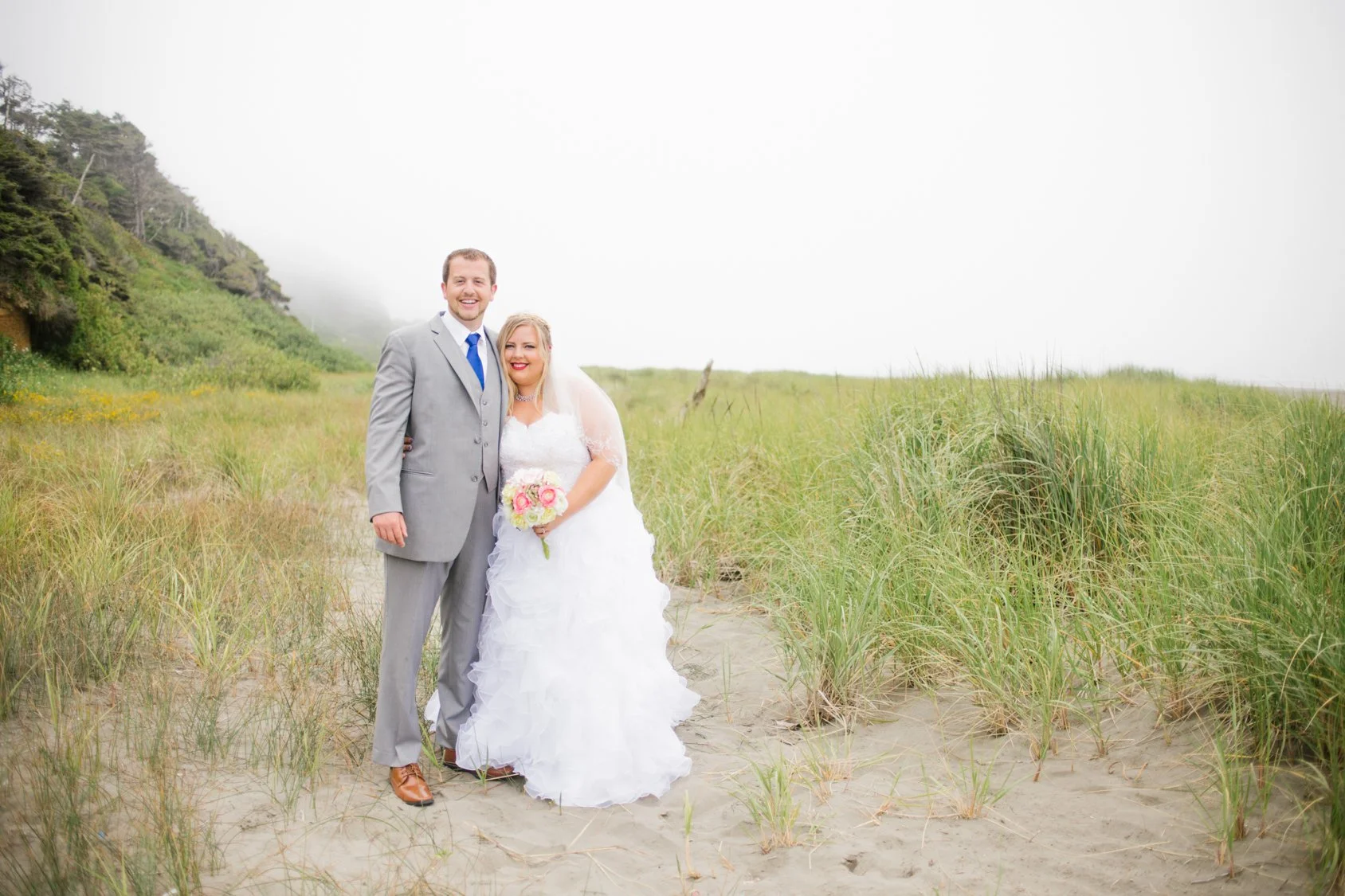 A newlywed couple standing together on a sandy beach with grass and green hills in the background. The groom in a grey suit and blue tie, and the bride in a white wedding gown holding a bouquet of pink and white flowers, smiling at the camera.