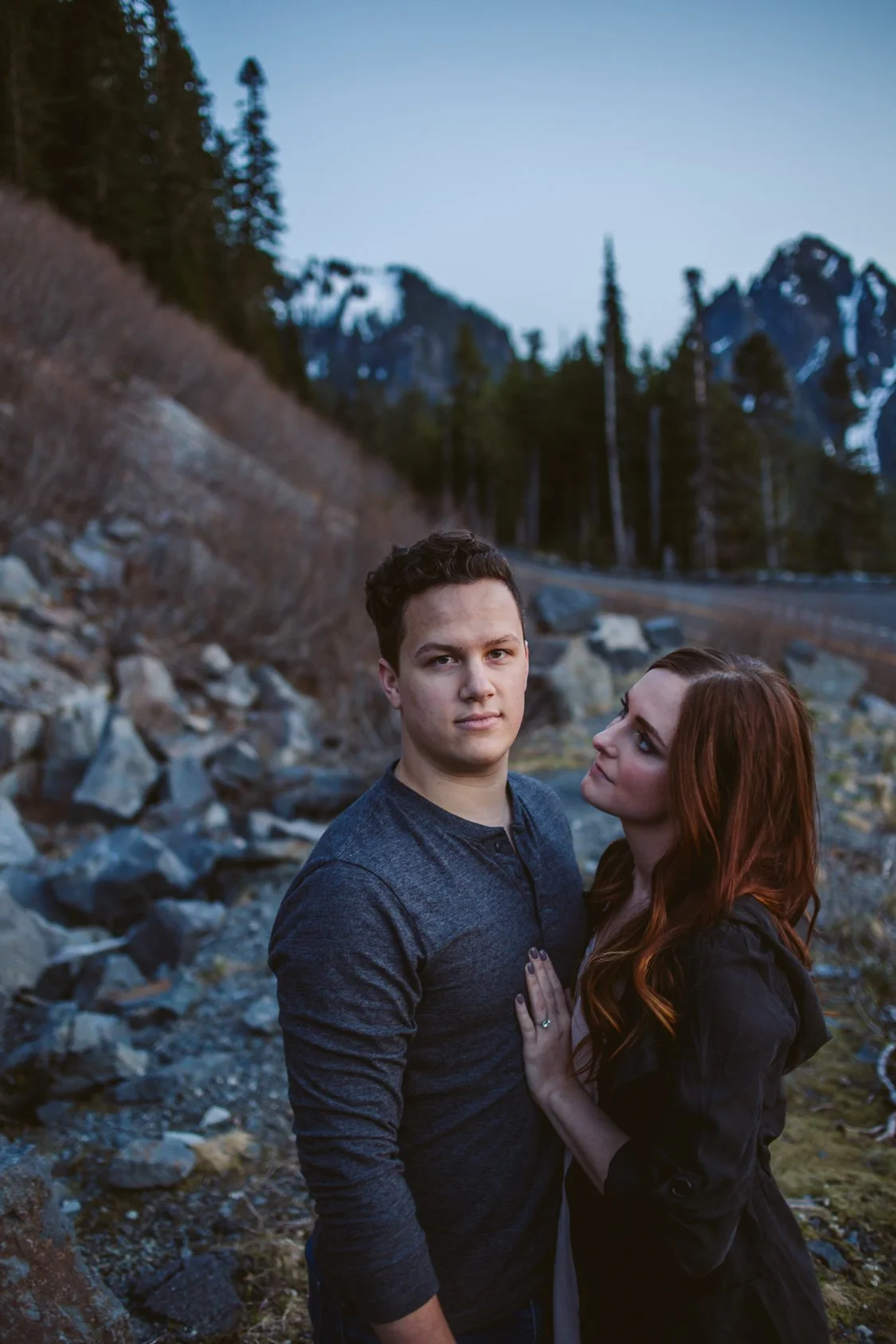A young man and woman stand close together outdoors in a rocky area, with a mountain range and pine trees in the background, during twilight.