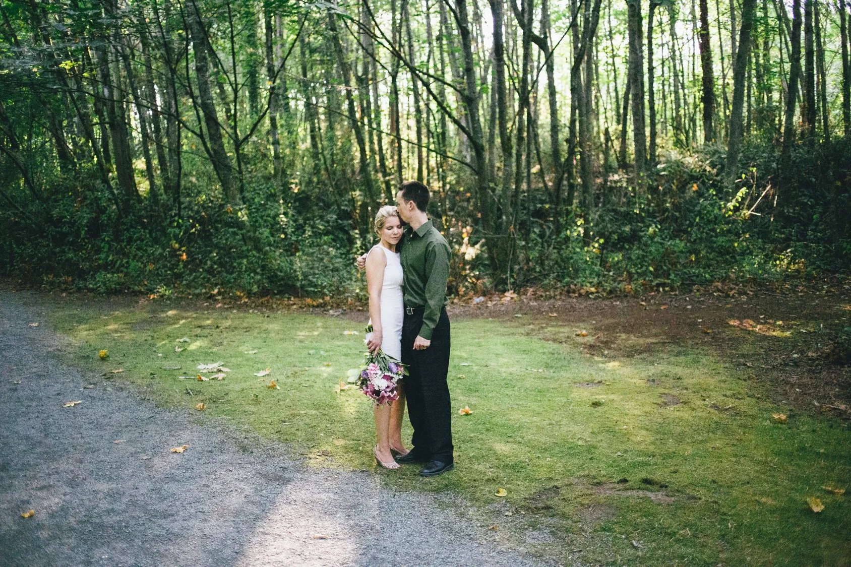 A couple standing close together in a forest, the woman in a white dress holding a bouquet and the man in a green shirt and black pants leaning his forehead against her temple.