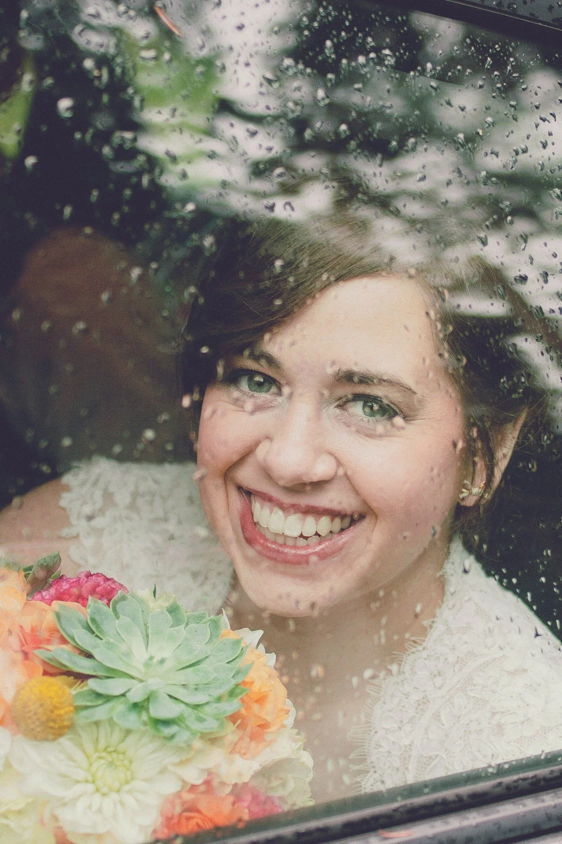 A woman smiling with visible teeth through a rain-covered window, holding a bouquet of colorful flowers.