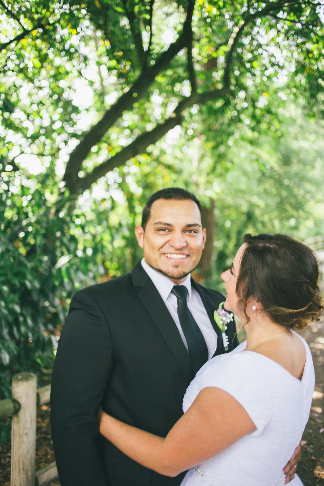 A smiling man in a black suit and tie standing beside a woman in a white dress, with their arms around each other in an outdoor setting with green trees in the background.