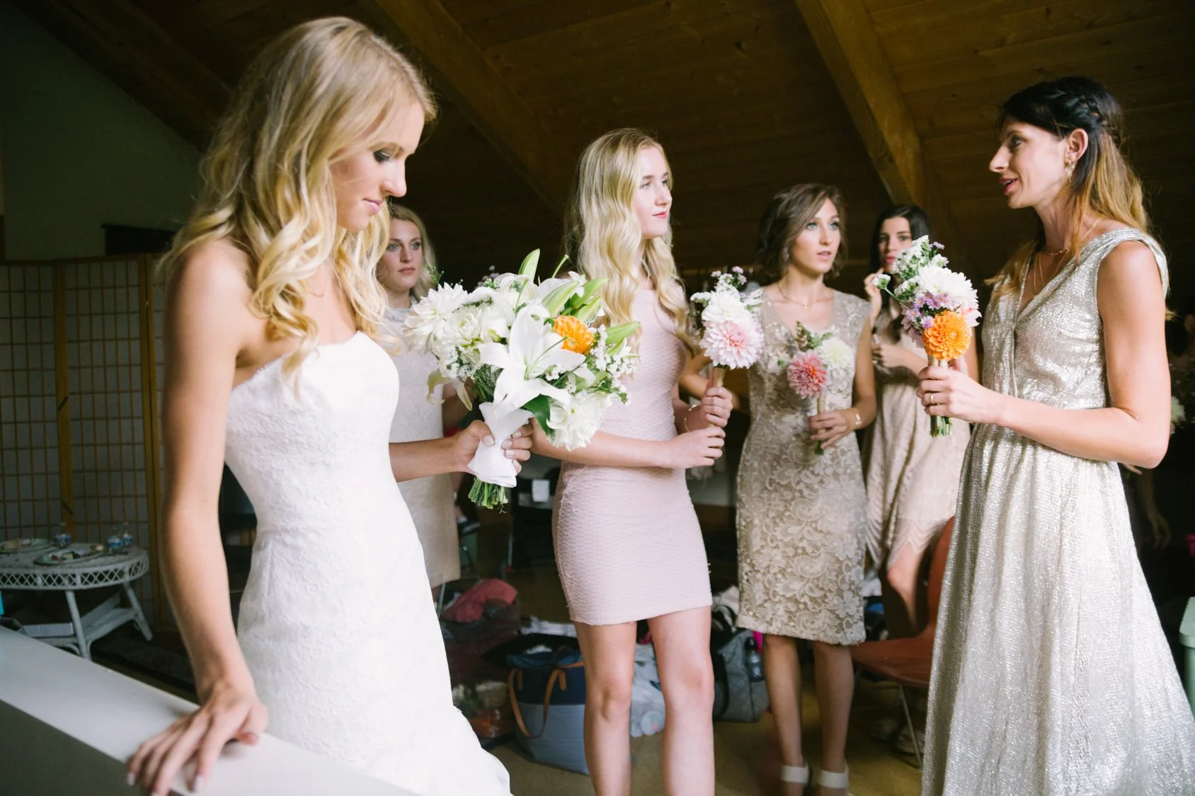 A group of women at a wedding, with one woman in a white wedding dress holding a bouquet of white flowers, and the others holding colorful flower arrangements, gathered indoors under a wooden ceiling.